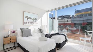 A modern bedroom with two beds, a nightstand, a chair, and a large window offering a city view.