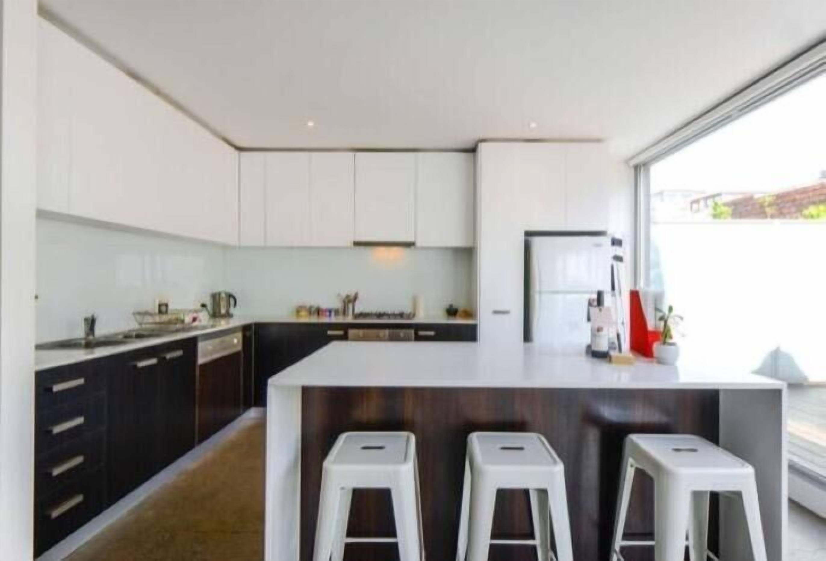 A modern kitchen with white cabinets, a central island with stools, and a large window providing natural light.