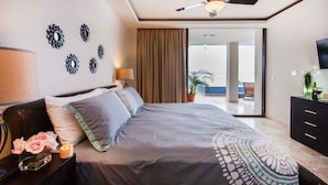 Un dormitorio con cama, ventilador de techo, mesita de noche con lámpara, decoración en la pared y vistas al balcón exterior.