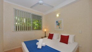 Habitación de hotel con cama, ventilador de techo, ventana con barrotes, un cuadro en la pared y mesita de noche con lámpara.