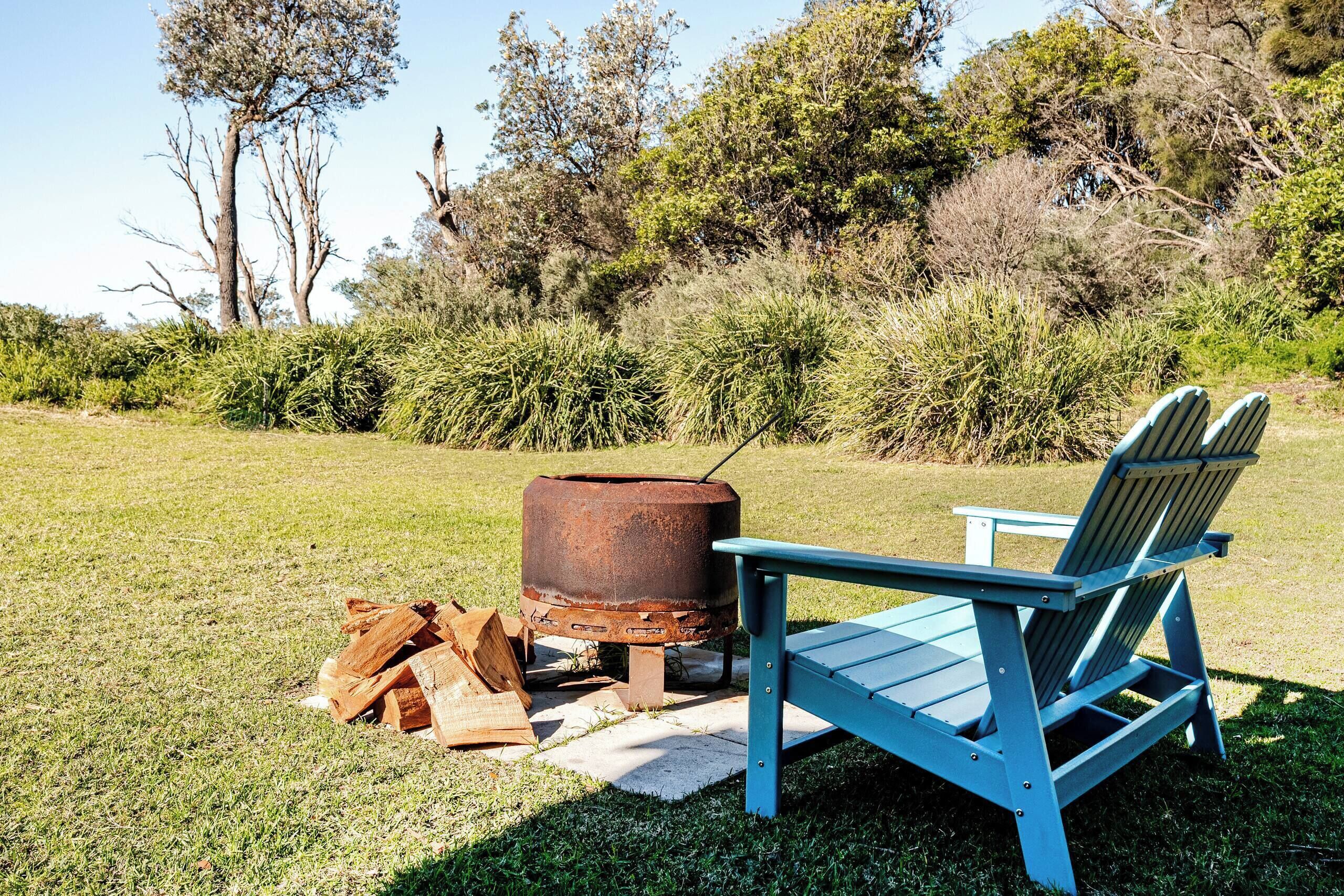 A blue Adirondack chair next to a metal fire pit with logs, set in a grassy outdoor area with trees and bushes in the background.