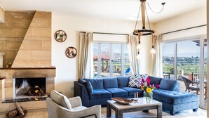 A living room with a fireplace, a blue sofa, a coffee table, and a view of the outdoors. - VILLA DEMETRA (4 ensuite bedrooms, BBQ, Wi-Fi, Prv. Swim. Pool, stunning view) (Argaka)