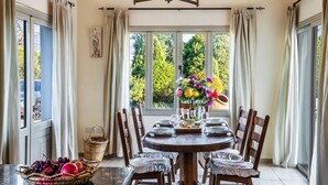 A dining area with a round table set for six, surrounded by wooden chairs. A vase with flowers is placed on the table, and a basket of fruit is on the floor beside the table. - VILLA DEMETRA (4 ensuite bedrooms, BBQ, Wi-Fi, Prv. Swim. Pool, stunning view) (Argaka)