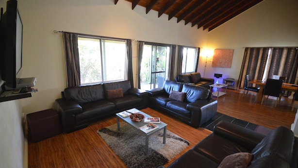 Una sala de estar con un conjunto de sofá, una mesa de centro y un televisor. Hay ventanas con cortinas, una lámpara y un cuadro en la pared.