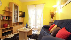 Un salon comprenant un canapé, une table basse, une bibliothèque, un téléviseur et une fenêtre avec des rideaux.
