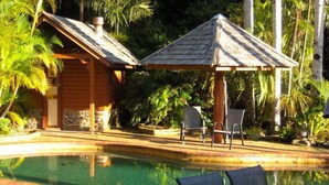 Uma área de piscina com um edifício de madeira e um gazebo, cercada por palmeiras.