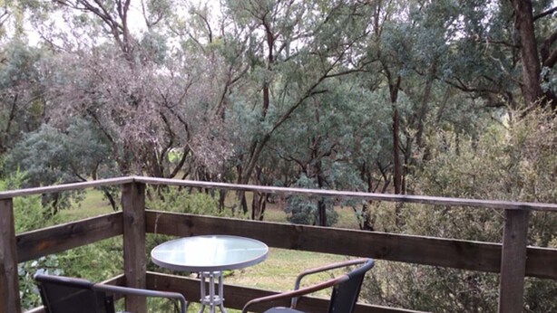 A wooden deck with a glass table and two chairs, overlooking a wooded area. - Frogsong Strawbale Cottage BLUE | Mansfield (Mountain Bay)