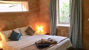 A hotel room with a bed, pillows, a lamp, and a window with curtains. - Frogsong Strawbale Cottage BLUE | Mansfield (Mountain Bay)