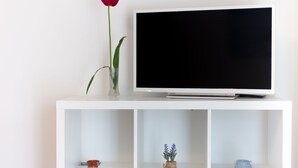 Un meuble de rangement blanc pour appareils électroniques, avec un téléviseur à écran plat, un vase contenant une fleur rouge et deux petits vases avec des plantes.