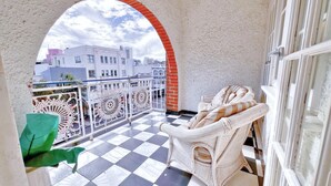 A balcony with a checkered floor, white wicker furniture, and a view of buildings through an archway.