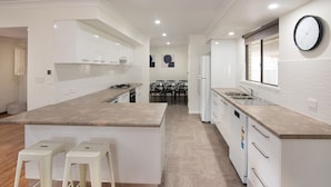 A modern kitchen with white cabinetry, a central island with stools, a built-in oven, and a clock on the wall. - SHELLEBRATIONS - Endless Summer (West Busselton)