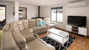 A modern living room with a large L-shaped beige leather sofa, a flat-screen TV, a white coffee table, and a black and white patterned rug. - SHELLEBRATIONS - Endless Summer (West Busselton)