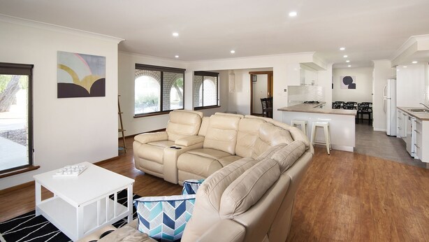 A modern living room with a beige sectional sofa, a small white table, and a patterned cushion. The room has wooden flooring, a kitchen area with white cabinets, and a dining area with black chairs. - SHELLEBRATIONS - Endless Summer (West Busselton)