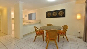 A dining area with a round table and wicker chairs, a kitchen in the background, and a wall-mounted artwork.