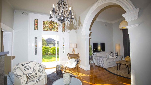 A living room with a chandelier, a white sofa, a wicker chair, and a television in the background. - Camellia Cottage - Beautiful 3 bedroom cottage with gorgeous gardens - stroll to local cafes! (Cessnock)