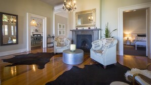 A spacious living room with a fireplace, two white armchairs, a chandelier, and a large mirror above the mantel. - Camellia Cottage - Beautiful 3 bedroom cottage with gorgeous gardens - stroll to local cafes! (Cessnock)
