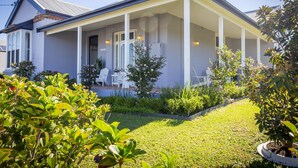 A two-story house with a covered porch, a lawn, and various plants. - Camellia Cottage - Beautiful 3 bedroom cottage with gorgeous gardens - stroll to local cafes! (Cessnock)