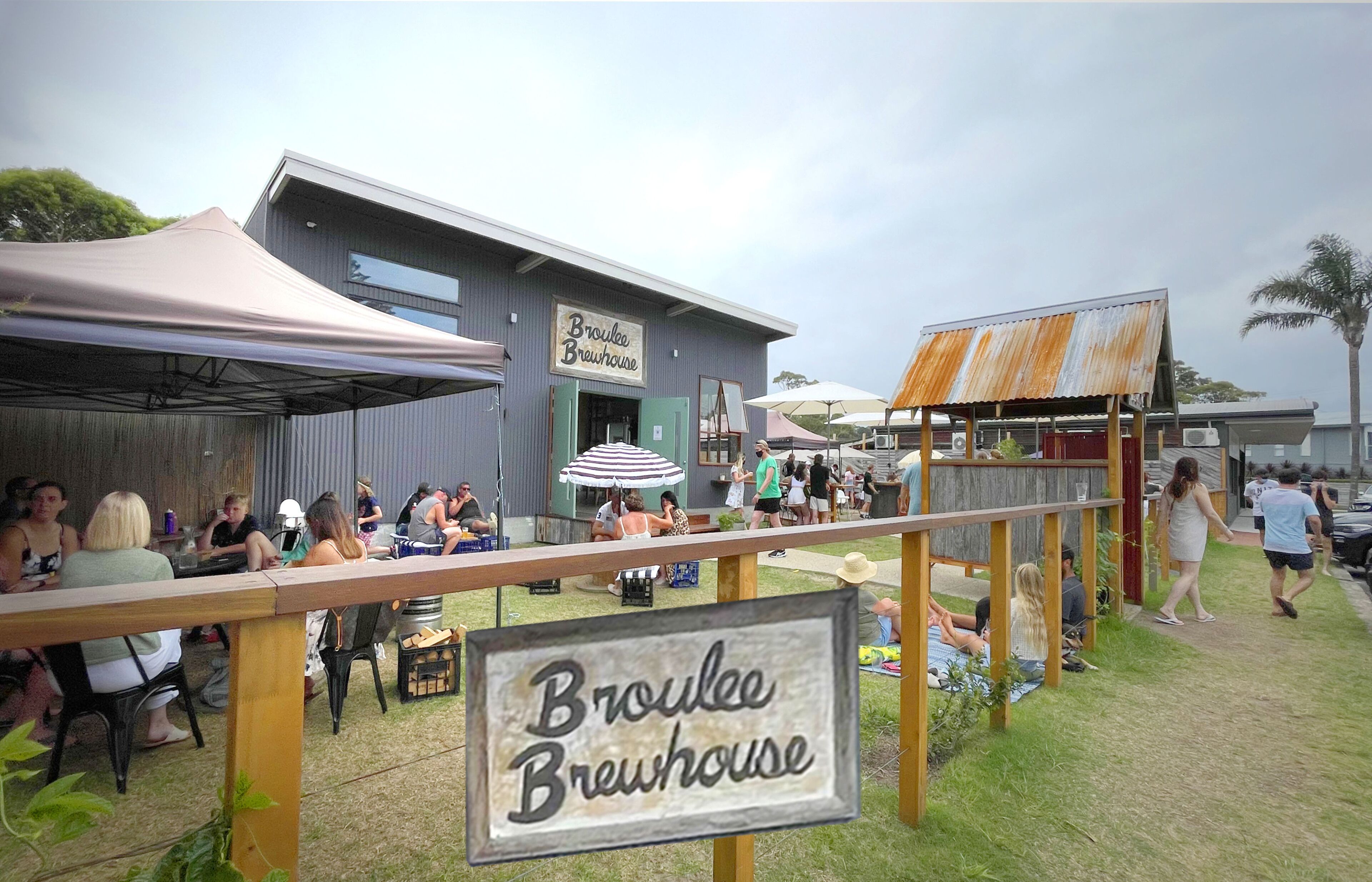 Ein Außenbereich mit Tischen und Stühlen, ein Schild für Broulee Brewhouse und ein rustikales Gebäude im Hintergrund.