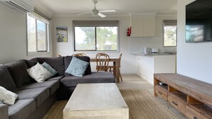 A modern living room with a sofa, coffee table, dining table, and chairs. There is a ceiling fan, large windows, and a kitchen area with a TV. - Self contained Top Unit - Surfs Up - Hear the sea at night. (Broulee)