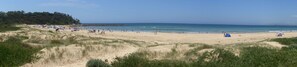 Une plage avec des tentes et des gens, des dunes de sable, et une zone boisée en arrière-plan.