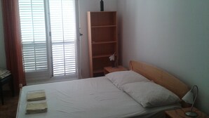 A bedroom with a bed, a nightstand, a wooden shelf, and a window with blinds. - Cosy apartments surrounded with green space (Petrovac)