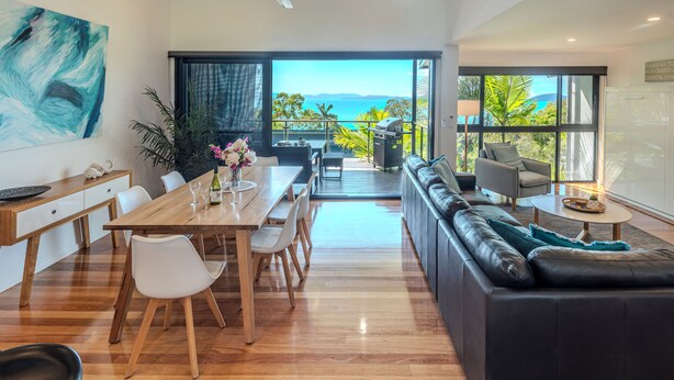 A modern living room with a wooden dining table, white chairs, a black leather sofa, and a view of the ocean through large windows. - Pinnacle 3, Seaview Apartment on Hamilton Island (Hamilton Island)