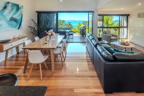 A modern living room with a wooden dining table, white chairs, a black leather sofa, and a view of the ocean through large windows.
