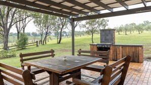 An outdoor patio with a wooden table and benches, a brick barbecue grill, and a view of a grassy field with trees. - The Cottage @ Hunter Gleann - Much more than the average cottage! (Pokolbin)