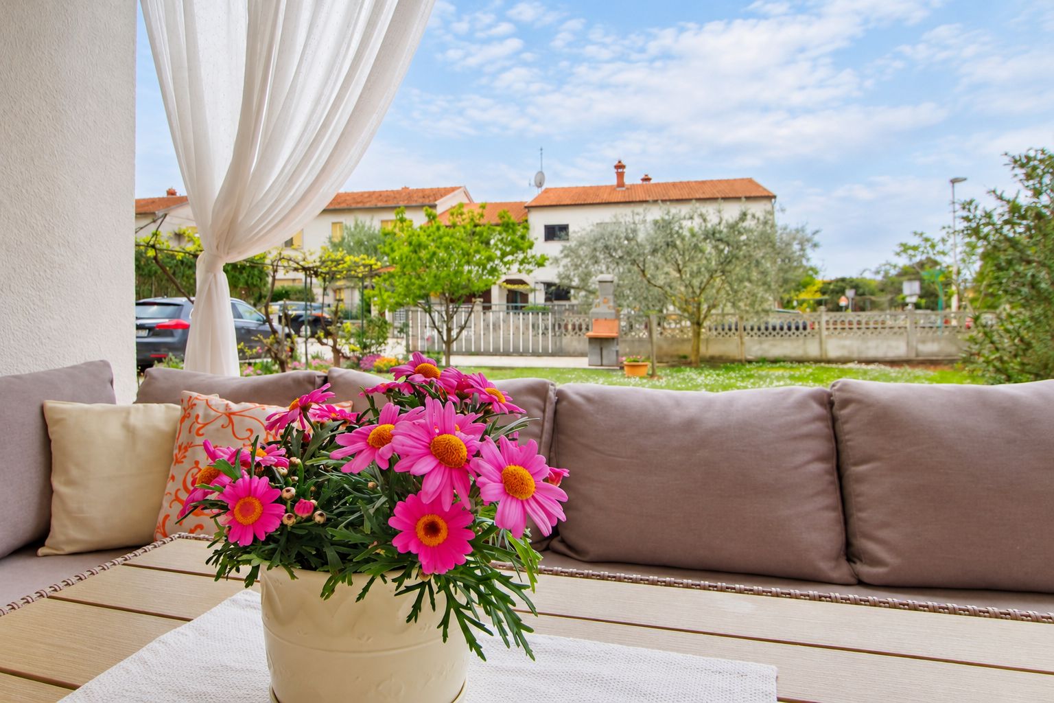 A beige sofa with a potted plant of pink flowers, a white curtain, and a view of a residential area with houses and trees.
