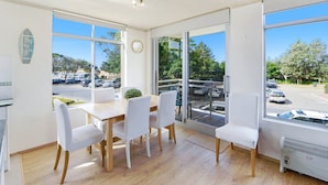 A bright room with a wooden table and white chairs, large windows with a view of a parking lot and trees. - Seaview 4 - Beautiful Views & Linen Supplied (Avoca Beach)
