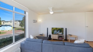 A modern living room with a grey sofa, a flat-screen TV, a ceiling fan, and a view of the street outside. - Seaview 4 - Beautiful Views & Linen Supplied (Avoca Beach)
