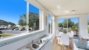 A modern kitchen with a large window, a sink, and a dining area with chairs and a table. - Seaview 4 - Beautiful Views & Linen Supplied (Avoca Beach)