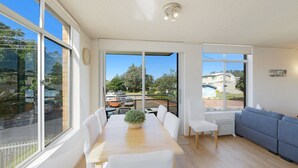 A bright, sunlit room with a dining table, chairs, and a sofa. Large windows offer views of a parking area and buildings. - Seaview 4 - Beautiful Views & Linen Supplied (Avoca Beach)