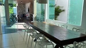 Un vaste espace dédié à la salle à manger, avec une longue table et des chaises blanches, de grandes fenêtres et une vue sur l’extérieur.