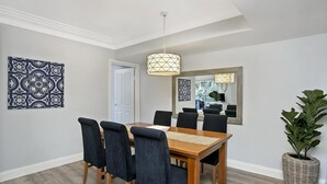 A dining area with a wooden table, six chairs, a hanging light fixture, a large mirror, and a potted plant.