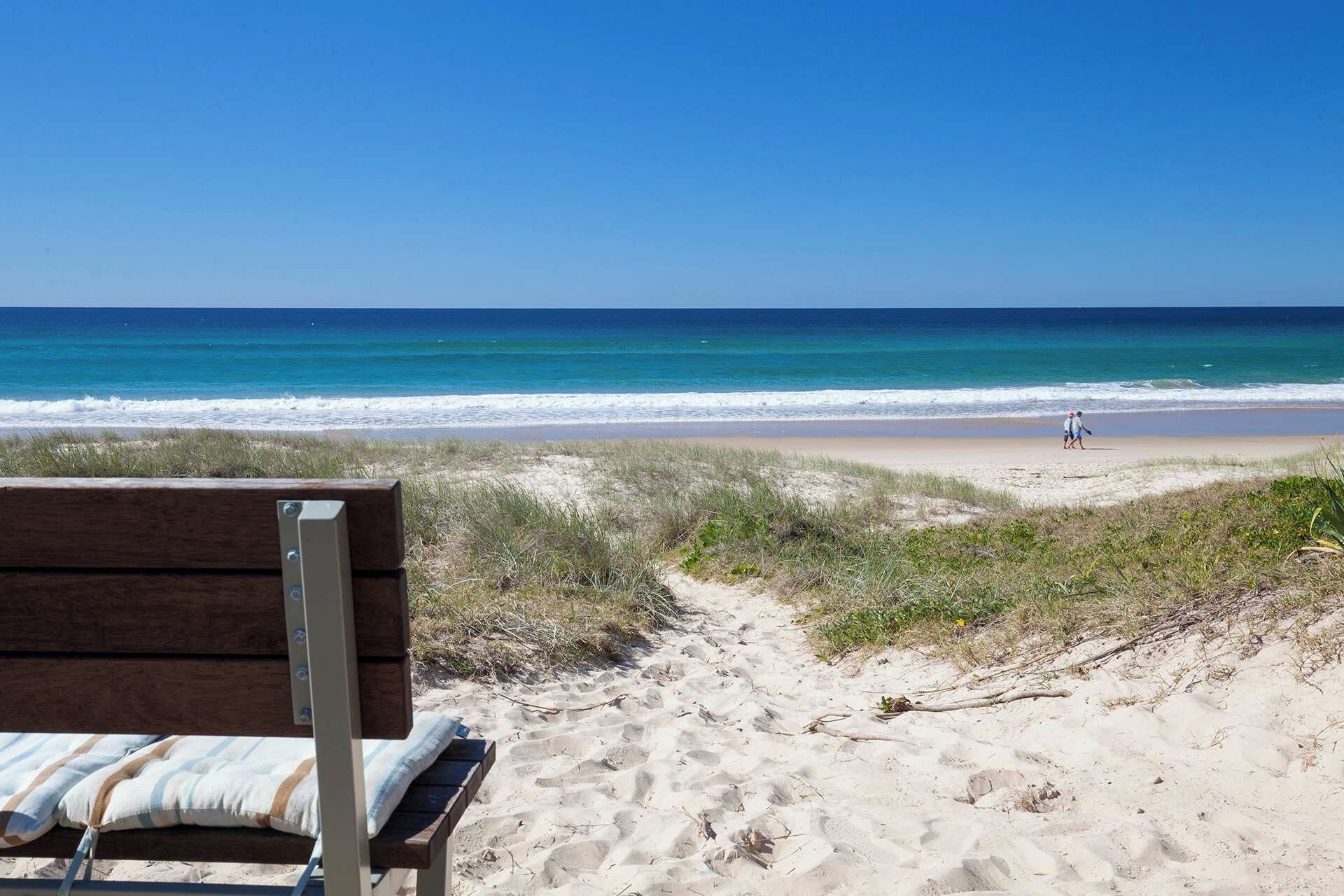 A beach view with a wooden bench and a path leading to the shore.
