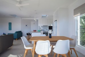 Eine moderne Küche mit weißen Schränken, ein Essbereich mit einem Holztisch und weißen Stühlen sowie ein großes Fenster mit Grünpflanzen draußen.