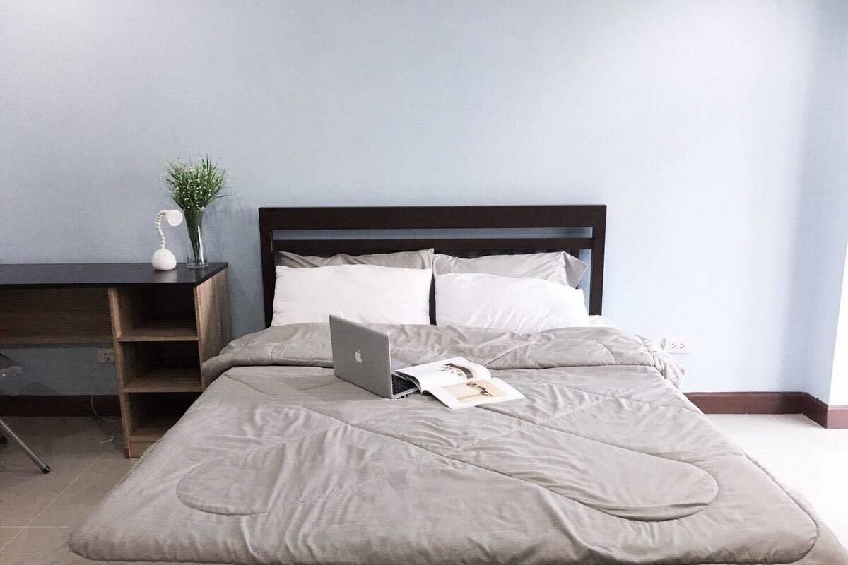 A neatly made bed with a laptop and a magazine, a dark wooden headboard, and a small desk with a plant and a telephone.