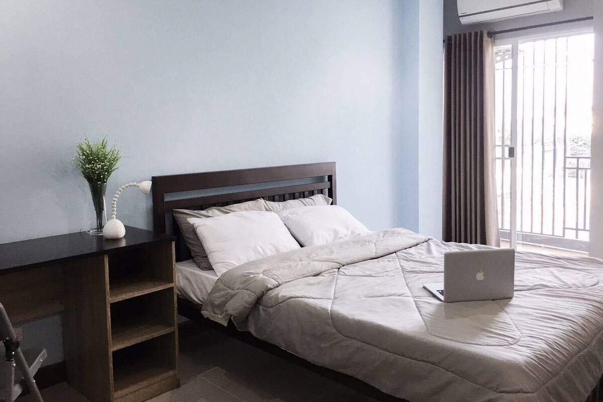 A bedroom with a bed, a desk, a laptop, a plant, and an air conditioning unit.