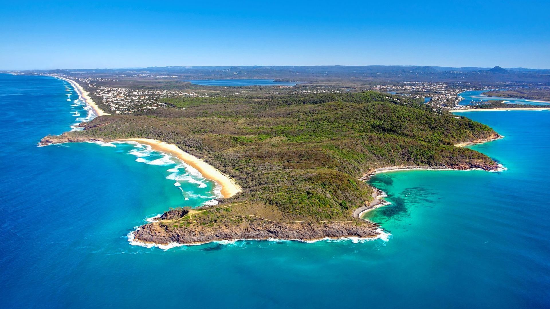 Ein Luftbild einer Küstenlandschaft mit Strand, bewaldetem Gelände und einem klaren, blauen Meer.