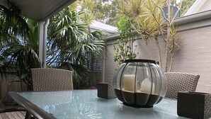 Un tavolo con piano in vetro, una candela in un vaso di vetro, sedie in vimini e piante in vaso in una zona patio all'aperto.