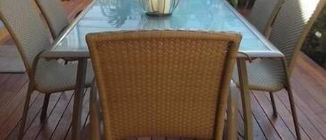 Une table à plateau en verre, entourée de chaises en osier, sur une terrasse en bois.