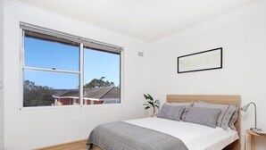 A bedroom with a large window, a bed with a grey blanket, a bedside table with a lamp, and a framed picture on the wall. - Mosman BOT01 Botanic Rd-Balmoral 2BR Balcony Steps from Balmoral Beach Esplanade (Mosman)