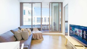 A modern living room with a sofa, a TV, and large windows offering a view of the outside. - Fawkner Residence 1B1B Condo with Pool&Gym Access (Melbourne)