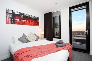 A hotel room with a bed, a red blanket, a gray pillow, a white pillow, a gray pillow, a red abstract painting, a window with curtains, and a view of a cityscape.