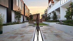 A modern building with a central water feature, surrounded by greenery and a clear sky.