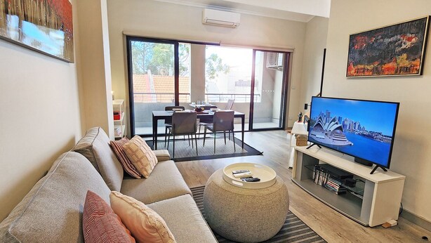 Ein Wohnzimmer mit einer Couch, einem Hocker und einem Flachbildfernseher. Es gibt einen Balkon mit Tisch und Stühlen.