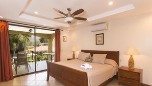 A bedroom with a bed, a ceiling fan, a nightstand with a lamp, and a sliding glass door leading to an outdoor area with a table and chairs. - Second-Floor Pool-View Condo – Your Perfect Beach Retreat in Jacó (Jacó)
