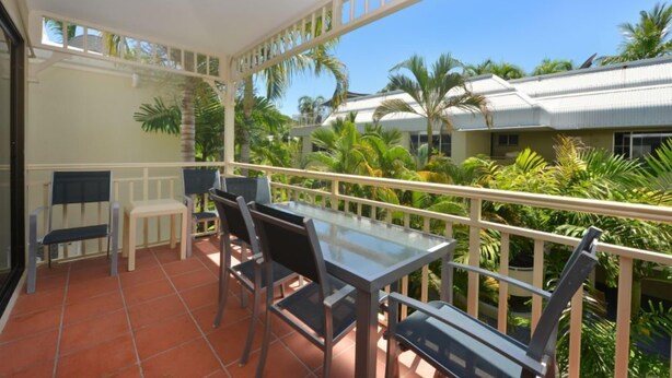 A balcony with a glass table and chairs, surrounded by a white railing, overlooking a lush garden with palm trees. - Driftwood Mantaray Three Bedroom Loft (Douglas)