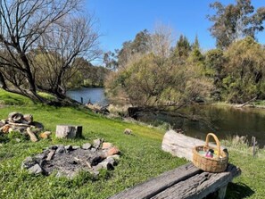 A picnic setup by a river with a wooden bench, a basket, and a fire pit.